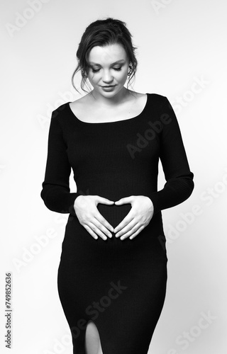 Black and white portrait of pregnant female in black dress with hands near pregnant belly with heart sign.