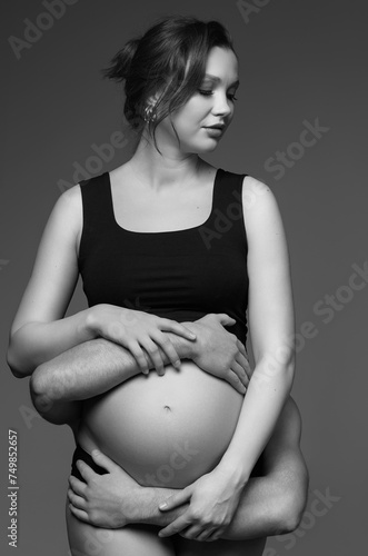 Black and white photo of man behind female and hugs her pregnant exposed belly.
