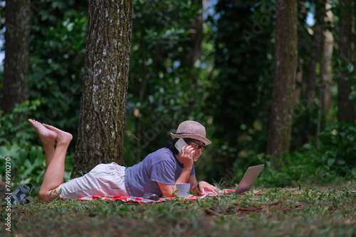 Woman enjoying her free time lying on the grass under shady trees in the park, making a phone call while working on her computer photo