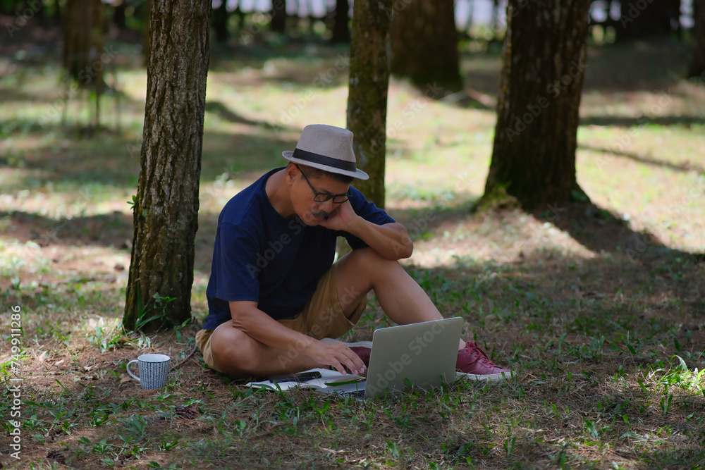 Man sitting on the grass under a shady pine tree in the forest while working on his laptop computer Man sitting on the grass under a shady pine tree in the forest while working on his laptop computer