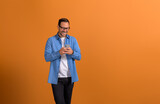 Cheerful male professional in glasses messaging over smart phone and standing on orange background