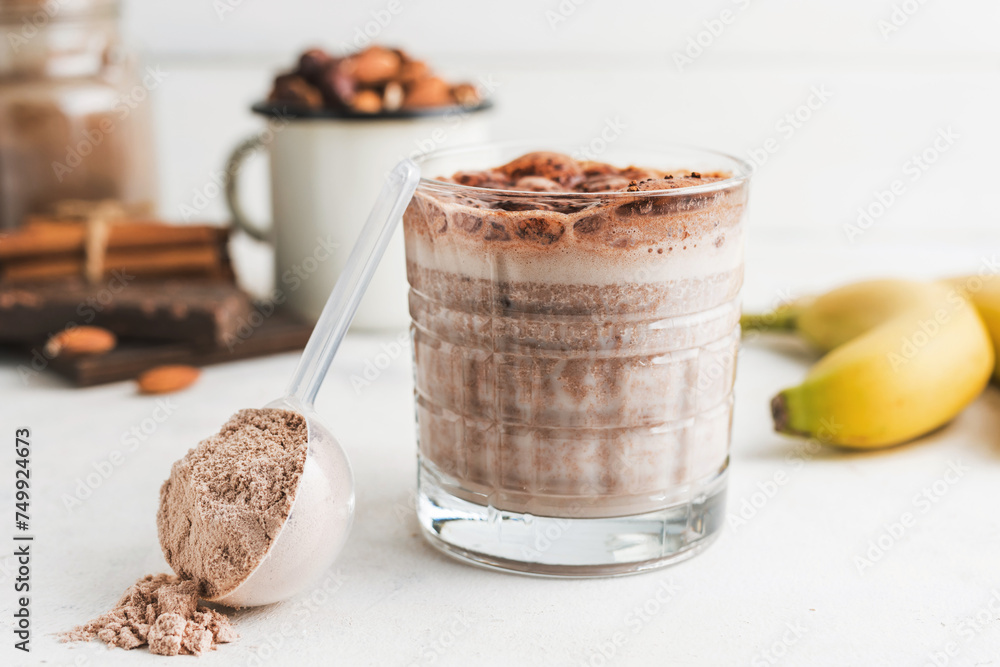 Glass with chocolate protein drink, milkshake smoothie on white table with bananas, protein powder in measuring spoon, protein bar, almond nuts and cinnamon sticks Glass with chocolate protein drink, milkshake smoothie on white table with bananas, protein powder in measuring spoon, protein bar, almond nuts and cinnamon sticks