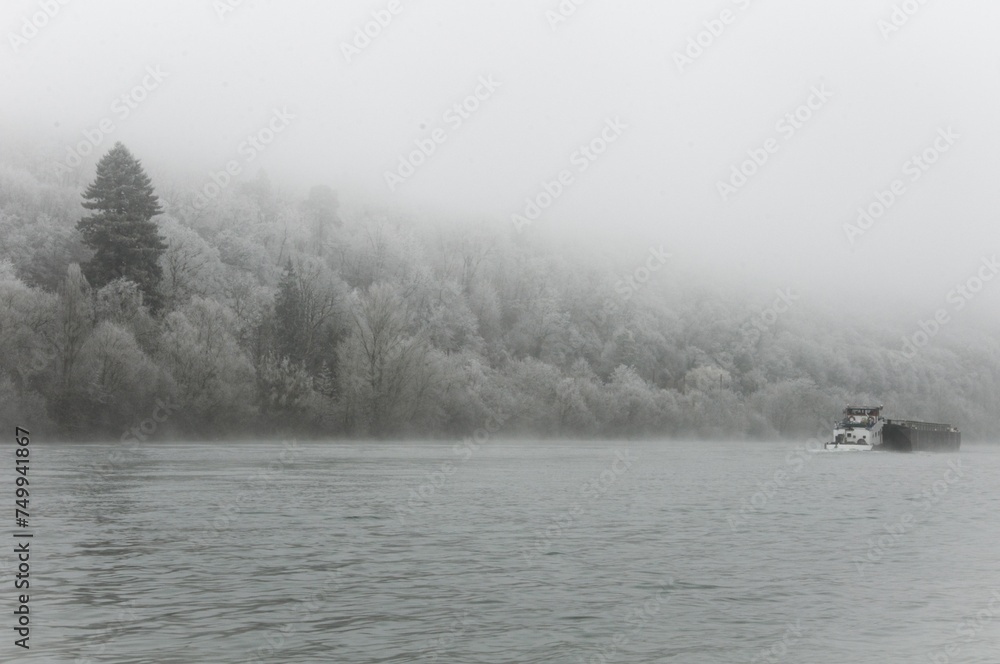 péniche avançant sur un cours d'eau au bord d'une forêt gelée, le tout dans le brouillard de l'hiver