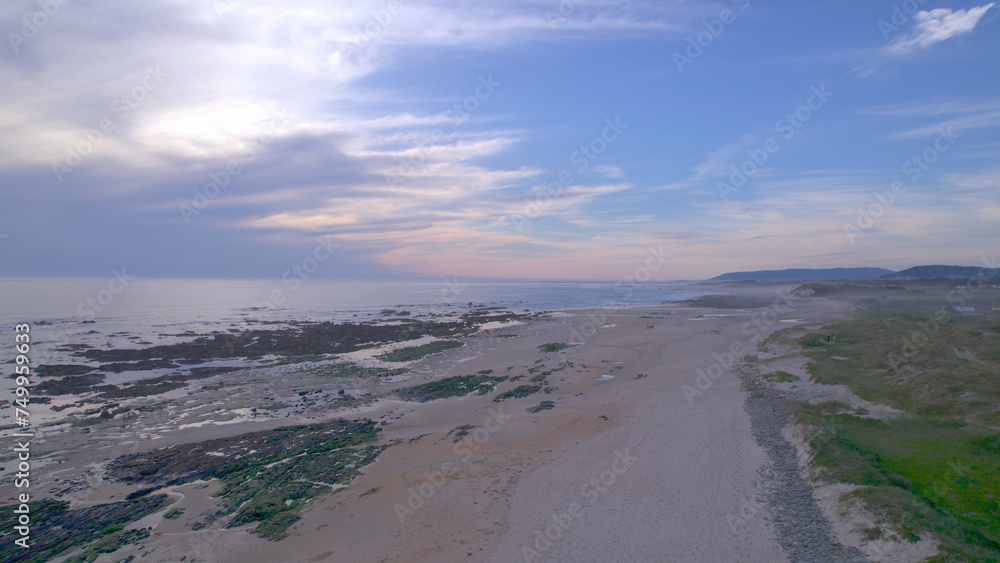Sea, beach boulders, pebble shore, and waves at sunset. Sea waves breaking on rocks. Beach with sand dunes and dusk sky in Rio de Moinhos, Esposende, Portugal.