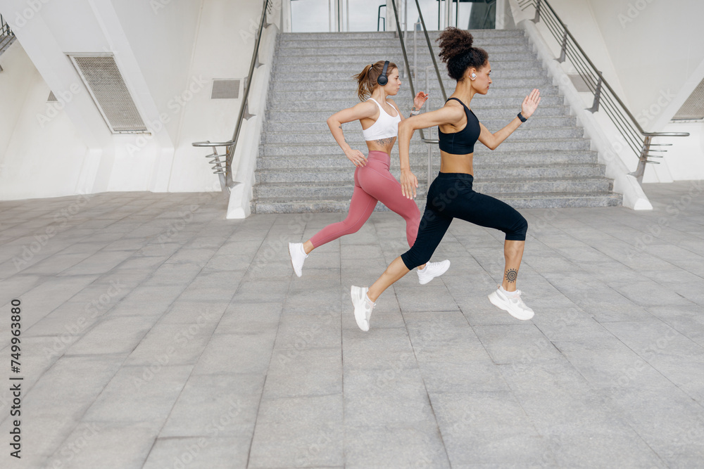 Side view of active women running side by side along an outdoor track on modern buildings background