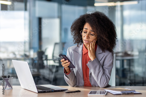 Upset young African American woman sitting in the office at the desk, leaning her head on her hand, holding the phone in her hand and looking bored at the screen.