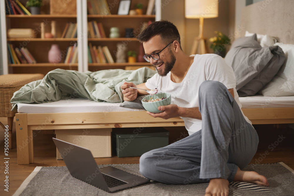 Young man with laptop having breakfast in bedroom Young man with laptop having breakfast in bedroom