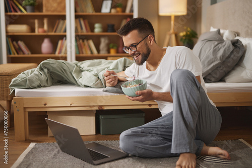 Young man with laptop having breakfast in bedroom