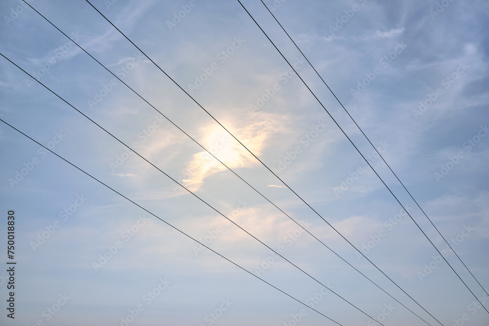 Diagonal hanging high-voltage transmission power lines against blue sky with clouds and shining sun. Empty space. Above-ground electric grid simple background. 