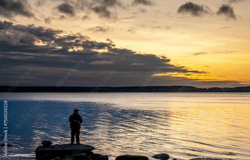 A fisherman with a fishing rod on the shore of the lake in the early morning.