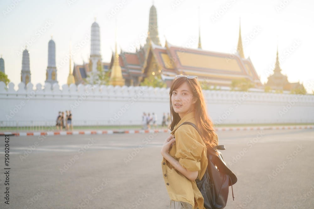 Fototapeta premium Traveler asian woman in her 30s, backpack slung over her shoulder, explores the intricate details of Wat Pra Kaew with childlike wonder. Sunlight dances on the golden rooftops.