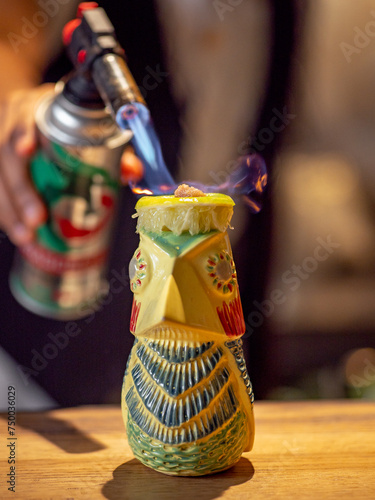 Bartender make tiki drink cocktail with gas torch. Barman burns brown sugar in tropical tiki cocktail on bar counter. Tiki cocktail in totem bird mug making process on bar in night club