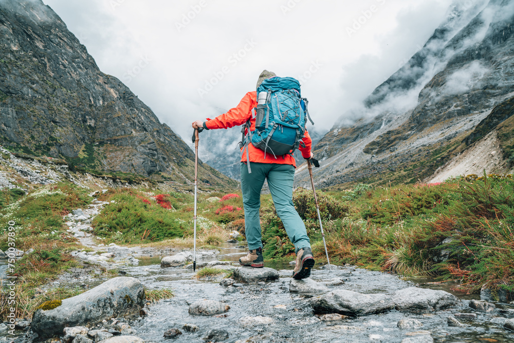 Moman with backpack and trekking poles crossing mountain creek during Makalu Barun National Park trek in Nepal. Mountain hiking and active people concept image.