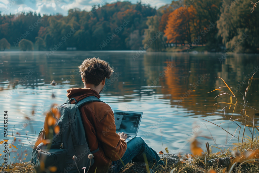 a young creative sitting by a lake and working on laptop