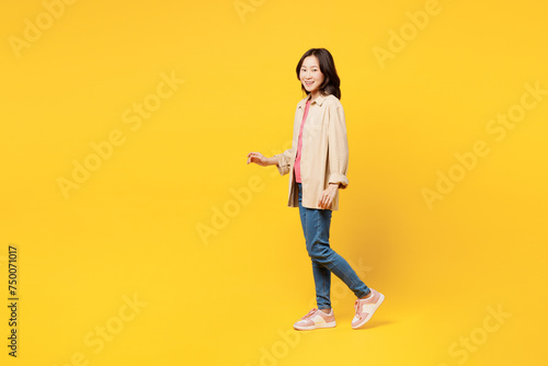 Full body side profile view young woman of Asian ethnicity she wearing pink t-shirt beige shirt pastel casual clothes walk go isolated on plain yellow background studio portrait. Lifestyle portrait.