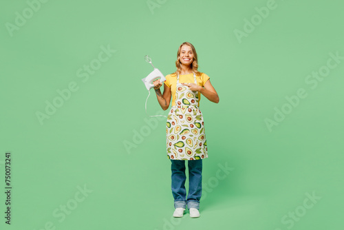 Full body young housewife housekeeper chef cook baker woman wear apron yellow t-shirt hold in hand point index finger on mixer looking camera isolated on plain green background. Cooking food concept.