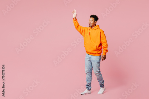 Full body side profile view young man of African American ethnicity he wears yellow hoody casual clothes walk go waving hand isolated on plain pastel light pink background studio. Lifestyle concept.
