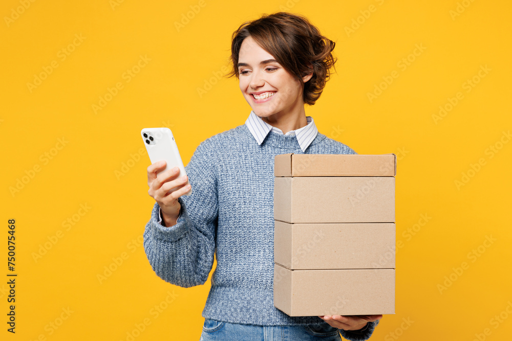 Φωτογραφία Young smiling woman she wears grey knitted sweater shirt casual clothes hold stack cardboard blank boxes use mobile cell phone isolated on plain yellow background studio portrait