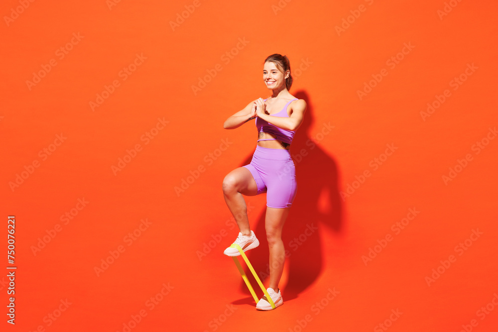 Full body fun young fitness trainer instructor woman sportsman wear purple top clothes in home gym hold elastic rubber band for legs isolated on plain orange background. Workout sport fit abs concept. Full body fun young fitness trainer instructor woman sportsman wear purple top clothes in home gym hold elastic rubber band for legs isolated on plain orange background. Workout sport fit abs concept.