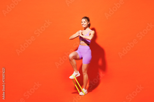 Full body fun young fitness trainer instructor woman sportsman wear purple top clothes in home gym hold elastic rubber band for legs isolated on plain orange background. Workout sport fit abs concept.