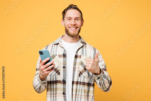 Young fun smiling happy Caucasian man he wear brown shirt casual clothes hold in hand use mobile cell phone show thumb up isolated on plain yellow orange background studio portrait. Lifestyle concept.