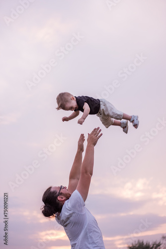 Billede på lærred Young, diversified father throws little son into air against backdrop of soft pastel sky at sunset