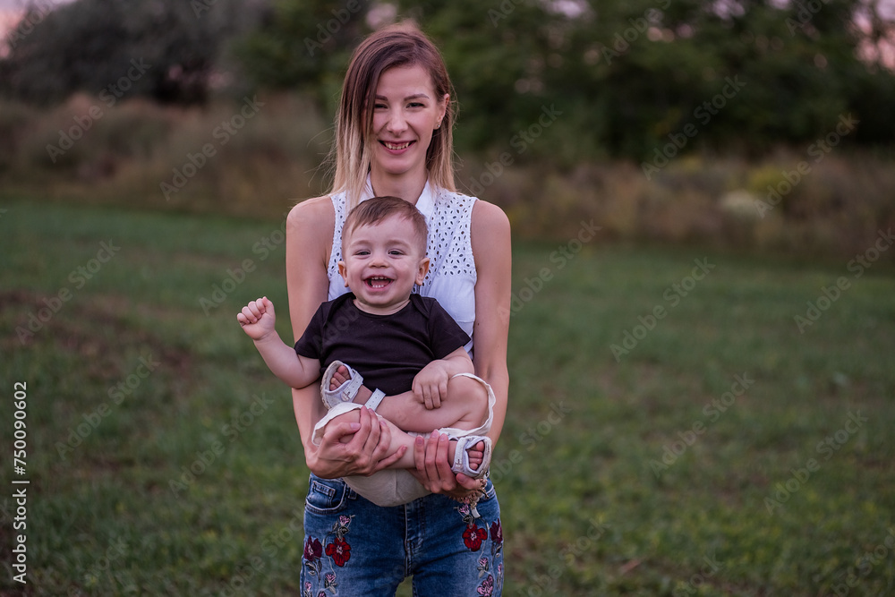 Fototapeta premium Young woman smiles, holds toddler son in her arms, standing in lush field with the soft glow of evening light. The concept of tactility, maternal connection with the child.