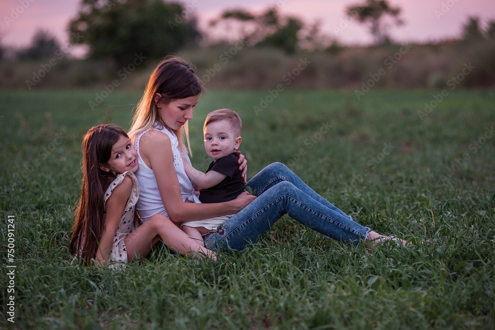 Mother embraces son and daughter, sitting on grassy field as twilight settles. Moment of tranquil affection, connection. Serene Family Time at Dusk. Woman hugging kids in green meadow. Mothers day Mother embraces son and daughter, sitting on grassy field as twilight settles. Moment of tranquil affection, connection. Serene Family Time at Dusk. Woman hugging kids in green meadow. Mothers day