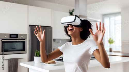 A laughing African American girl wearing virtual reality glasses in a modern kitchen, dressed in a white T-shirt, emotionally gesticulates with her hands while controlling the application. copy space photo