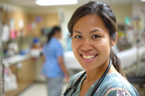 A smiling woman wearing hospital scrubs stands in a hospital room with a stethoscope up to The room is equipped with medical equipment and another employee in the background