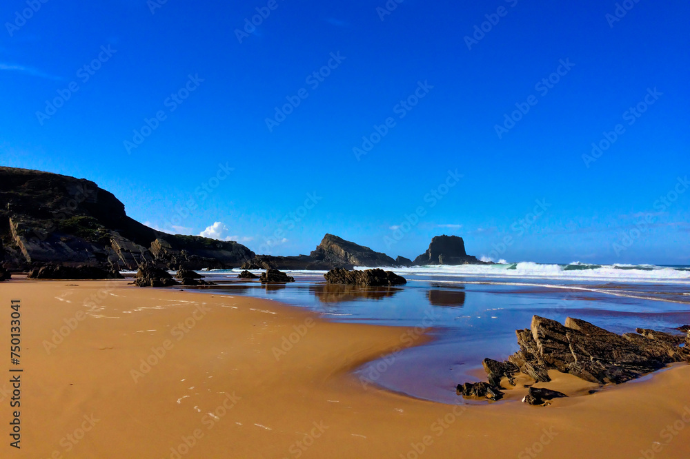 A sandy beach with rocky outcrops, waves gently crashing, under a clear blue sky.