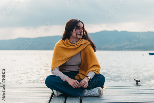 A young woman sits cross-legged on a wooden dock, wrapped in a yellow shawl, looking pensively across the waters of a mountain lake