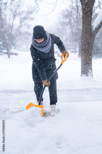 A young woman dressed in a winter coat, gloves, hat and scarf holding a yellow snow shovel, clearing a pathway after a blizzard