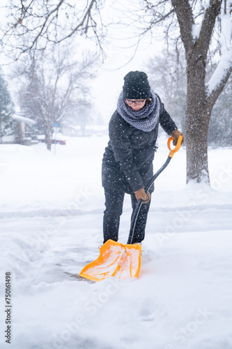 A young woman dressed in a winter coat, gloves, hat and scarf holding a yellow snow shovel, clearing a pathway after a blizzard