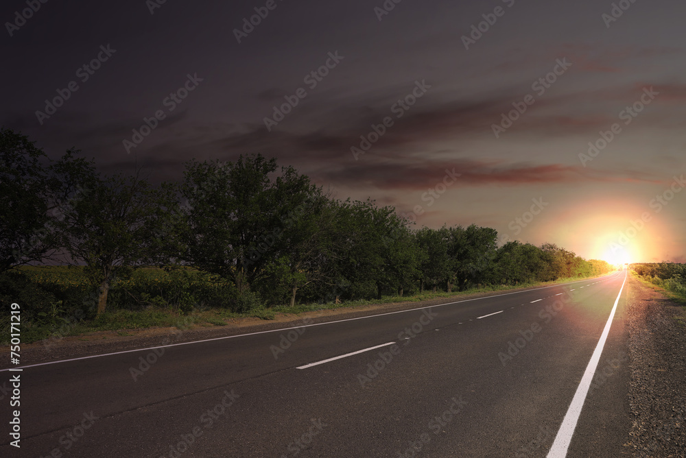 Empty asphalt road and trees under dark sky with rising sun