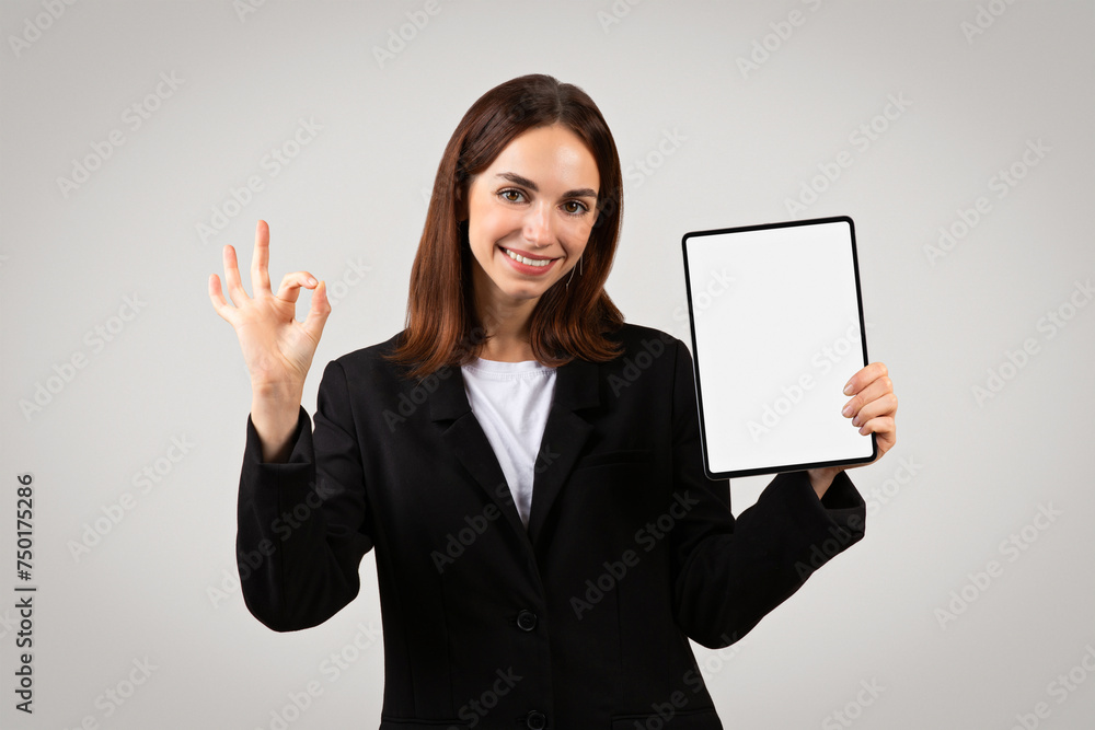 A poised young businesswoman holds a tablet with a blank screen in one hand while making an 'okay' A poised young businesswoman holds a tablet with a blank screen in one hand while making an 'okay'