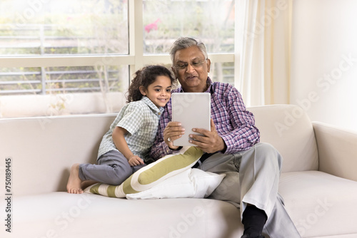 Positive Indian granddad showing online learning application to little grandkid holding tablet, sitting on home sofa, using digital gadget for online family communication, smiling, speaking