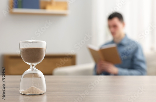 Hourglass with flowing sand on desk. Man reading book in room, selective focus