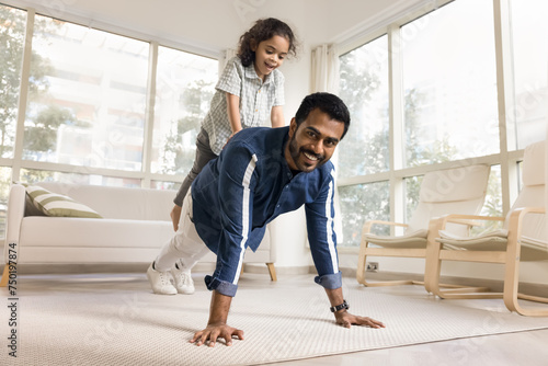 Happy strong athletic Indian dad piggybacking little daughter  keeping static yoga plank on floor  doing pushups with kids weight  looking at camera  exercising for strength  training body