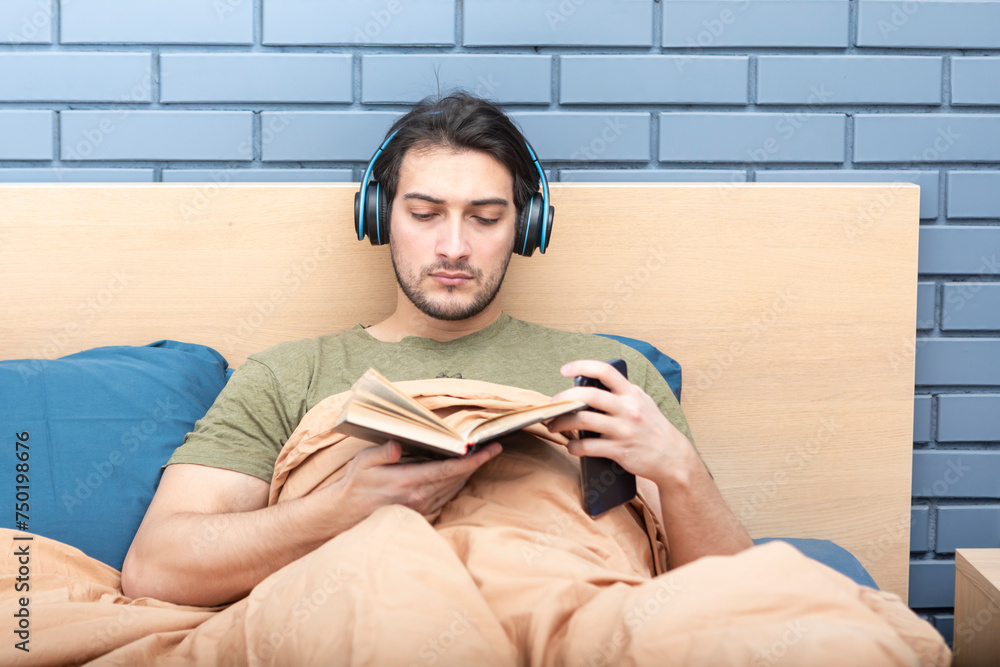 Obraz premium Young man lying at the bed, reading a book and listening audiobook at the same time