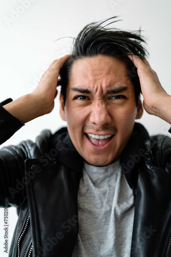 Latin man wearing black leather jacket standing against white wall looking stressed holding his head with his hands