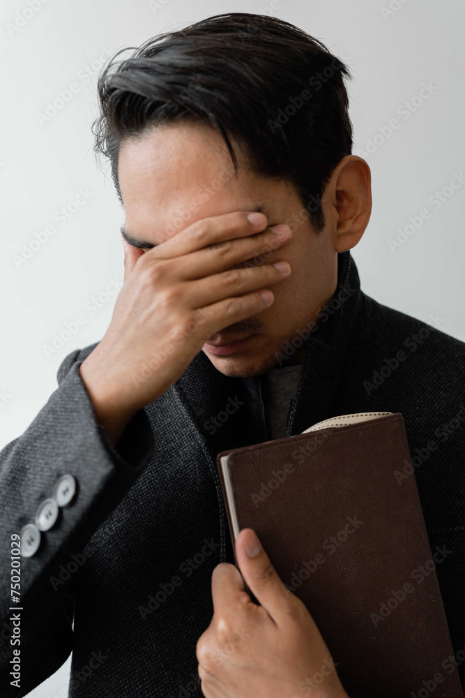 Latin man wearing a coat holding a book covering his eyes with a hand Latin man wearing a coat holding a book covering his eyes with a hand