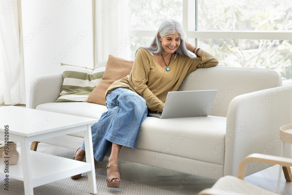 Happy senior elder Latin woman typing on laptop, relaxing on comfortable couch in white colored home interior, looking at screen, laughing, enjoying leisure in Internet, online communication Happy senior elder Latin woman typing on laptop, relaxing on comfortable couch in white colored home interior, looking at screen, laughing, enjoying leisure in Internet, online communication