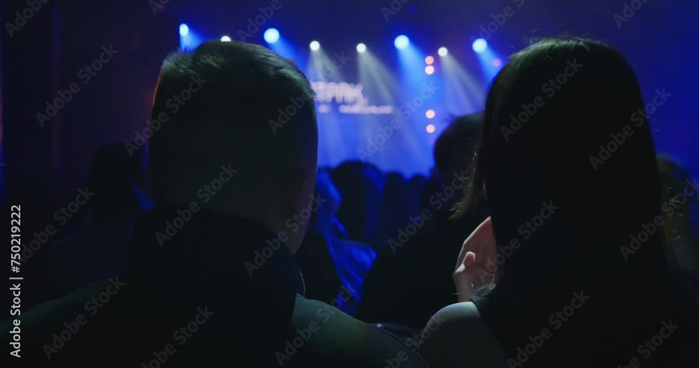 A young man and a millennial woman are standing at a concert in a dark hall and waiting for the artist to come on stage. Unrecognizable faces, a view from the auditorium to the stage. High quality 4k A young man and a millennial woman are standing at a concert in a dark hall and waiting for the artist to come on stage. Unrecognizable faces, a view from the auditorium to the stage. High quality 4k