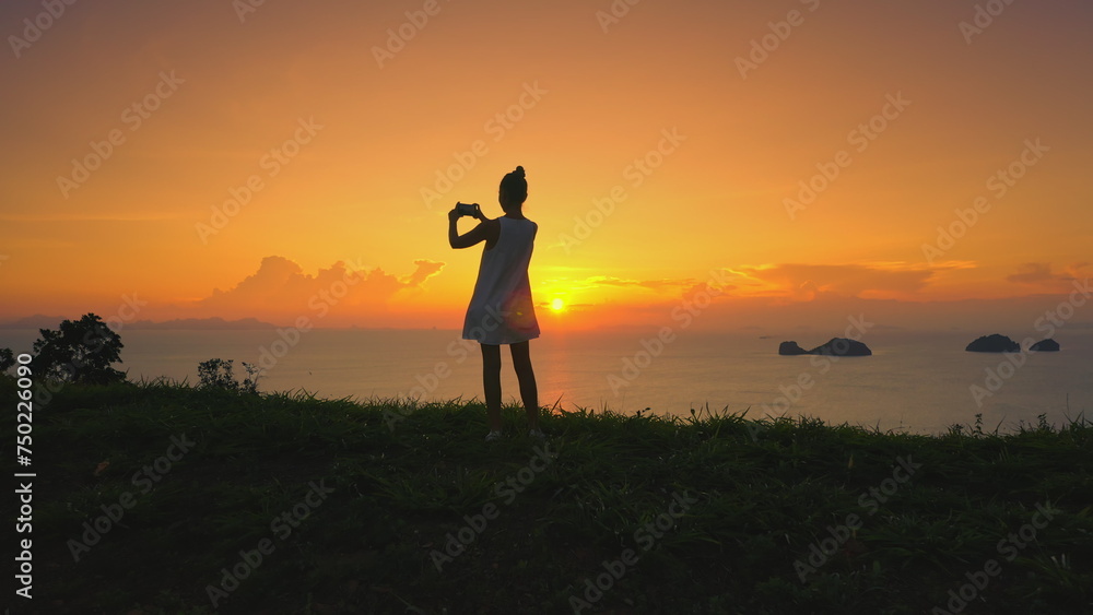 Woman silhouette against vibrant sunset, radiant glow setting sun. Grassy field, orange sky, girl with camera capture nature beauty ocean islands. Female tourist on summer vacation. Lifestyle holiday