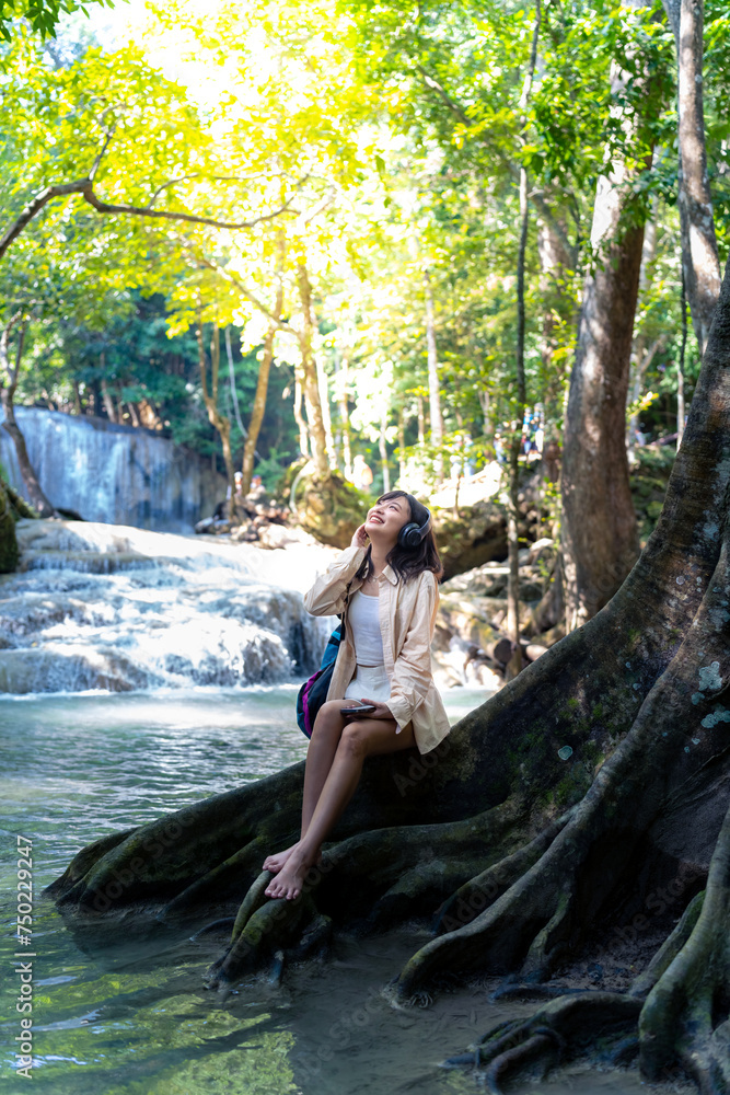 Fototapeta premium Young Asian woman relax and enjoy outdoor lifestyle travel nature tropical forest on summer holiday vacation. Happy generation z girl listening to the music on headphones and mobile phone by waterfall