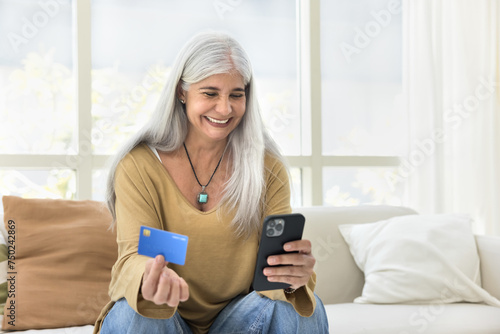 Cheerful grey haired senior Latin woman making money transaction with online financial service for payment, holding mobile phone, paying for Internet purchase, showing credit card