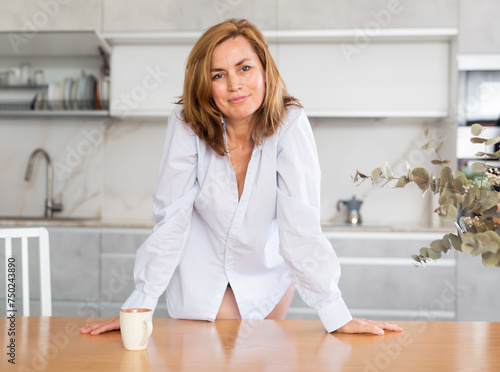 Delightful woman in only shirt over her naked body with cup of coffee in her hands sits on chair in the kitchen