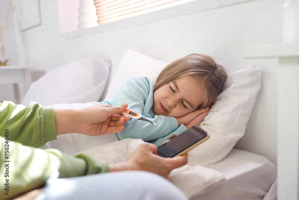 Sick little girl with her mother holding thermometer and mobile phone in bedroom Sick little girl with her mother holding thermometer and mobile phone in bedroom
