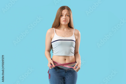 Upset young woman in tight jeans measuring her belly on blue background. Weight gain concept